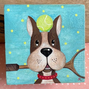 Puede incluir: Pintura cuadrada de madera de un perro con una pelota de tenis en la cabeza y una raqueta de tenis en la boca. El perro es marrón y blanco con un collar rojo. El fondo es azul claro con lunares amarillos.