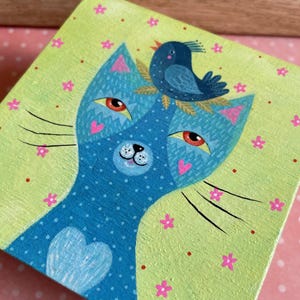 Puede incluir: Una obra de arte cuadrada pintada a mano que representa un gato azul con un pecho en forma de corazón y un pájaro azul posado en su cabeza. El gato tiene orejas rosas en forma de corazón y ojos rojos. El fondo es verde claro con flores rosas y puntos rojos.