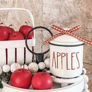 K&ouml;nnte beinhalten: Wei&szlig;e Keramikdose mit einem rot-wei&szlig; karierten Band um den Deckel. Die Dose hat das Wort "Apples" in Rot auf der Vorderseite. Die Dose steht auf einem wei&szlig;en, mehrst&ouml;ckigen Tablett mit roten &Auml;pfeln und wei&szlig;en Perlen.