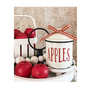 K&ouml;nnte beinhalten: Wei&szlig;e Keramikdose mit schwarzem Rand und roter Aufschrift "APPLES". Die Dose hat ein rot-wei&szlig;es Gingham-Band, das um den Deckel gebunden ist. Die Dose steht auf einem wei&szlig;en, mehrst&ouml;ckigen Tablett mit roten &Auml;pfeln.