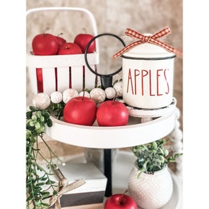 K&ouml;nnte beinhalten: Ein wei&szlig;er Etagere mit roten &Auml;pfeln, einem wei&szlig;en Keramiktopf mit rotem Deckel und dem Wort "APPLES" in roten Buchstaben und einem wei&szlig;en Keramiktopf mit einer gr&uuml;nen Pflanze.