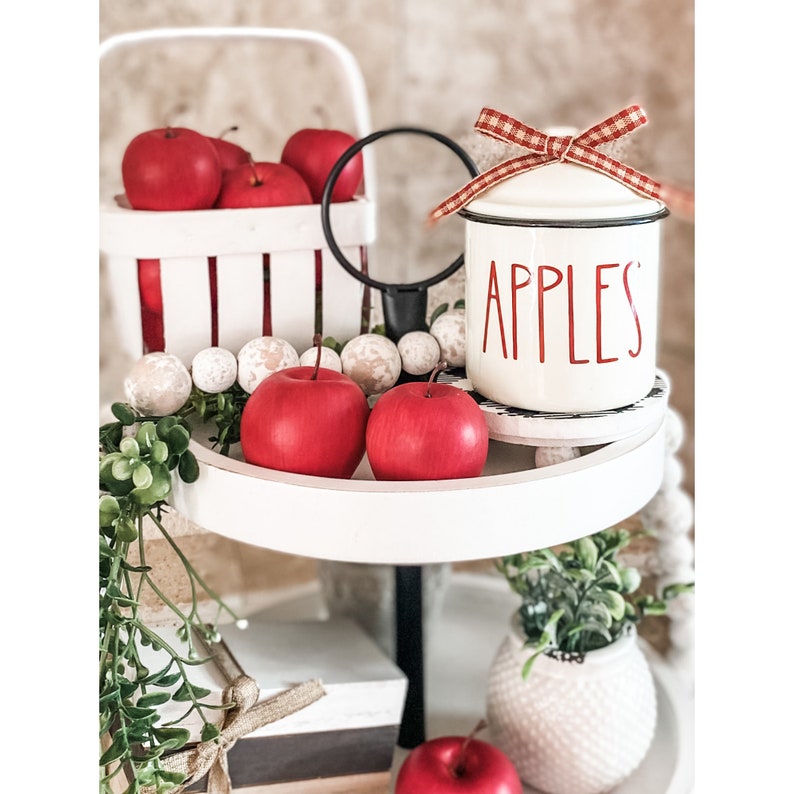 K&ouml;nnte beinhalten: Ein wei&szlig;er Etagere mit roten &Auml;pfeln, einem wei&szlig;en Keramiktopf mit rotem Deckel und dem Wort "APPLES" in roten Buchstaben und einem wei&szlig;en Keramiktopf mit einer gr&uuml;nen Pflanze.