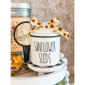 May include: White ceramic canister with a black rim and the text "Sunflower Seeds" in black lettering. The canister has a sunflower-patterned ribbon tied around the lid.