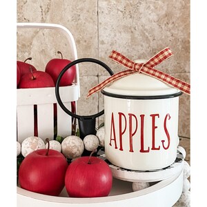 K&ouml;nnte beinhalten: Eine wei&szlig;e Emailledose mit schwarzem Rand und roter Aufschrift "APPLES". Die Dose hat ein rot-wei&szlig;es Gingham-Band, das um den Deckel gebunden ist. Die Dose ist von roten &Auml;pfeln und wei&szlig;en Perlen umgeben.
