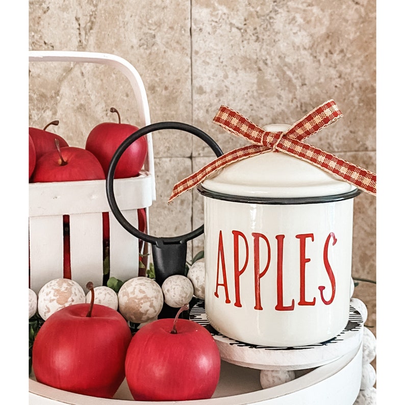 K&ouml;nnte beinhalten: Wei&szlig;e Emaille-Dose mit schwarzem Rand und roter Aufschrift "APPLES". Die Dose hat einen wei&szlig;en Deckel mit einem rot-wei&szlig; karierten Band, das zu einer Schleife gebunden ist.