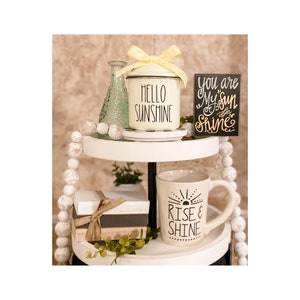 May include: A two-tiered white tiered tray with white beads, a green glass vase, a white enamel canister with a yellow bow and the text "Hello Sunshine", a white mug with black text that says "Rise & Shine", a small wooden sign with the text "You are my sun & I'll let you shine", and a wrapped gift box.
