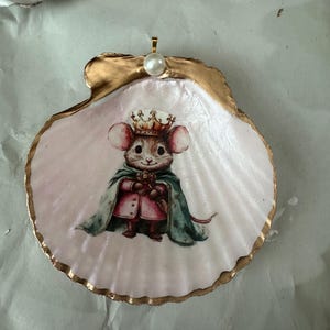 May include: A decorated scallop shell with a painted image of a mouse wearing a crown and cape. The shell has a gold-painted edge and a pearl attached at the top. The mouse is holding a small object.