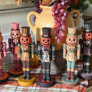May include: A collection of decorative Halloween nutcrackers. Each nutcracker has a pumpkin head, dressed in various outfits with top hats and bow ties. The color palette includes orange, pink, green, and black, with detailed facial expressions and clothing.