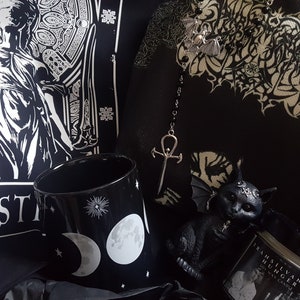 May include: A black and white gothic themed gift box with a black mug with a moon and stars design, a black cat figurine with wings, a silver ankh necklace, and a black candle with the text "Transylvanian Hunger" on the label.