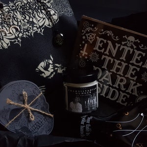 May include: A black box filled with gothic themed items, including a skull coaster, a candle with the text "Transylvanian Hunger Soy Wax Candle", a black pouch with a white floral design, and a black card with the text "Enter the Dark Side".
