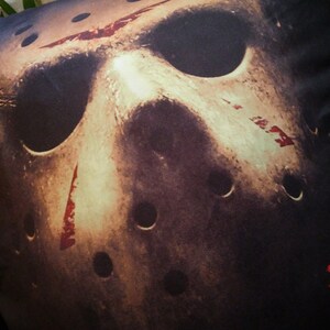 May include: A black pillow with a graphic of a hockey mask with red blood splatters. The text "FRIDAY THE 13TH" is printed in red on the bottom right corner of the pillow.