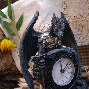May include: A black dragon figurine holding a clock with Roman numerals. The dragon is wearing silver armour and has wings spread out. The clock face is white with black hands.