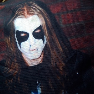 May include: A black and white photo of a person with long dark hair wearing black face paint with white accents. The person is wearing a black shirt and a black leather jacket. The background is a brick wall.