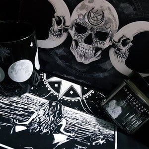 May include: Black and white illustration of a woman sitting on a beach with a crescent moon and stars in the sky. A black mug with a white crescent moon and stars design, a black and white skull with a crescent moon and star design, and a black candle jar with the text "Transylvanian Hunger" are also in the image.