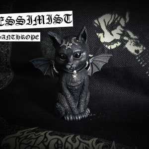 Peut inclure: Figurines de chat noir avec des ailes de chauve-souris, un pentagramme sur la tête et un collier argenté avec un pendentif pentagramme. La figurine est posée sur un fond en tissu noir. Le texte "PESSIMIST" et "MISANTHROPE" est visible au-dessus de la figurine.