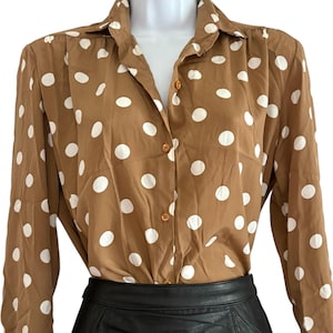 May include: A long-sleeved button-down shirt in a medium brown color featuring a repeating pattern of white polka dots. The shirt has a classic collar, a front button placket with matching brown buttons, and is tucked into a black skirt.