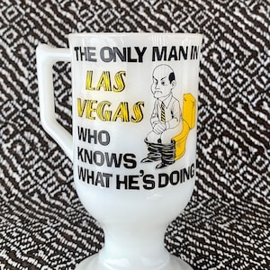 May include: White ceramic mug with a black and yellow design. The mug has a handle and a wide base. The text on the mug reads "The only man in Las Vegas who knows what he's doing". A cartoon man is sitting on a toilet.