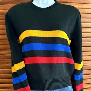 Pu&ograve; includere: Maglione nero con strisce orizzontali gialle, blu e rosse. Il maglione ha un girocollo e polsini e orlo a coste. Anche le maniche presentano il motivo a righe. Il maglione &egrave; esposto su un manichino.
