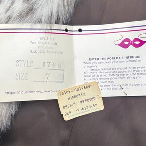 May include: A white rectangular clothing tag with a purple mask graphic and the text ENTER THE WORLD OF INTRIGUE. The tag lists material composition as 80% modacrylic, 20% acrylic, and 100% polypropylene back, with style 1780 and size 1 printed.