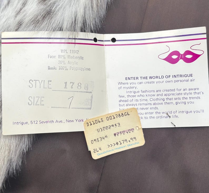 May include: A white rectangular clothing tag with a purple mask graphic and the text ENTER THE WORLD OF INTRIGUE. The tag lists material composition as 80% modacrylic, 20% acrylic, and 100% polypropylene back, with style 1780 and size 1 printed.