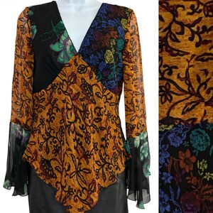 May include: A long-sleeved blouse featuring a V-neckline and a multi-panel design. The garment combines a mustard yellow fabric with a dark brown vine pattern and black panels adorned with a vibrant floral print in blue, green, and red tones.