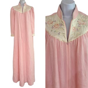 May include: A floor-length light pink robe featuring a high collar and a front zipper closure. The upper chest area has a cream-colored panel decorated with embroidered floral patterns in shades of pink, green, and purple, with matching cuffs.