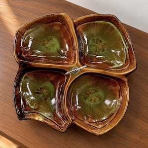 May include: A square ceramic serving dish with four rounded compartments, featuring a glossy glaze that transitions from deep brown at the edges to a translucent olive green in the center. A small silver-toned metal handle is centered.