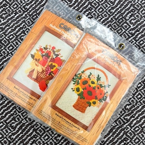May include: Two framed crewel embroidery kits. One features a basket of fruit, the other a basket of flowers. The frames are wood-toned, and the kits are sealed in plastic. The text on the kits reads "Basket of Fruit Crewel Kit" and "Basket of Flowers Crewel Kit".