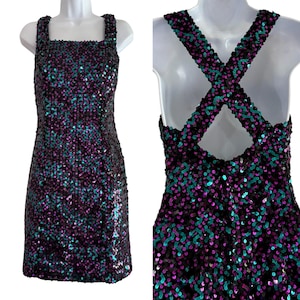 May include: A sleeveless mini dress covered in shimmering black, purple, and teal sequins. The front features a square neckline, while the back displays wide straps that cross over each other in an X-shape, leaving an open cutout design.