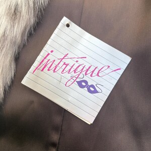 May include: A square white tag with horizontal black lines featuring the word Intrigue in pink cursive script and a purple masquerade mask icon. The tag has a single hole punch at the top center and is positioned against a dark brown fabric.