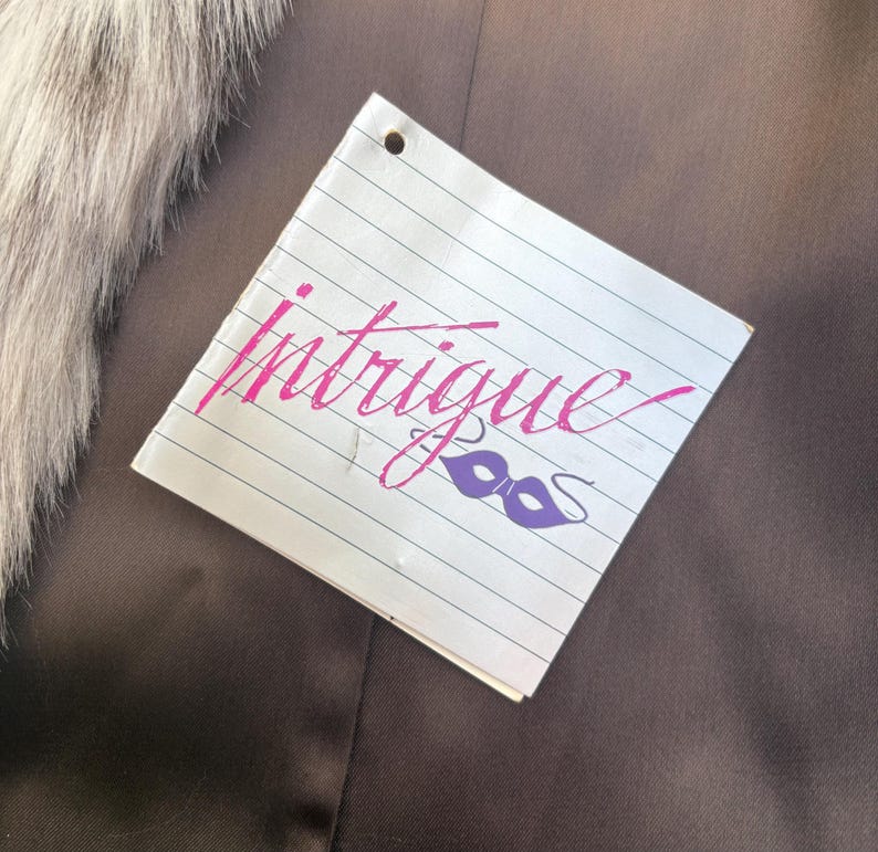 May include: A square white tag with horizontal black lines featuring the word Intrigue in pink cursive script and a purple masquerade mask icon. The tag has a single hole punch at the top center and is positioned against a dark brown fabric.