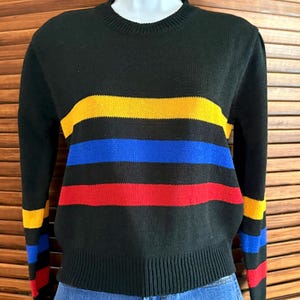 Pu&ograve; includere: Maglione nero con strisce orizzontali gialle, blu e rosse. Il maglione ha un girocollo a coste e polsini a coste. Le maniche presentano anche strisce negli stessi colori. Il maglione &egrave; esposto su un manichino.