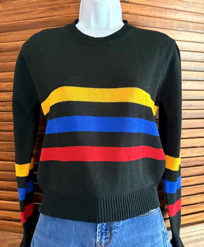 Pu&ograve; includere: Maglione nero con strisce orizzontali gialle, blu e rosse. Il maglione ha un girocollo a coste e polsini a coste. Le maniche presentano anche strisce negli stessi colori. Il maglione &egrave; esposto su un manichino.