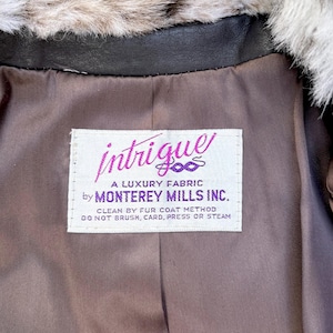 May include: A brown jacket interior with a white label reading intrigue A LUXURY FABRIC by MONTEREY MILLS INC. CLEAN BY FUR COAT METHOD DO NOT BRUSH, CARD, PRESS OR STEAM. The jacket features a dark brown lining and a fluffy grey and white faux fur collar.