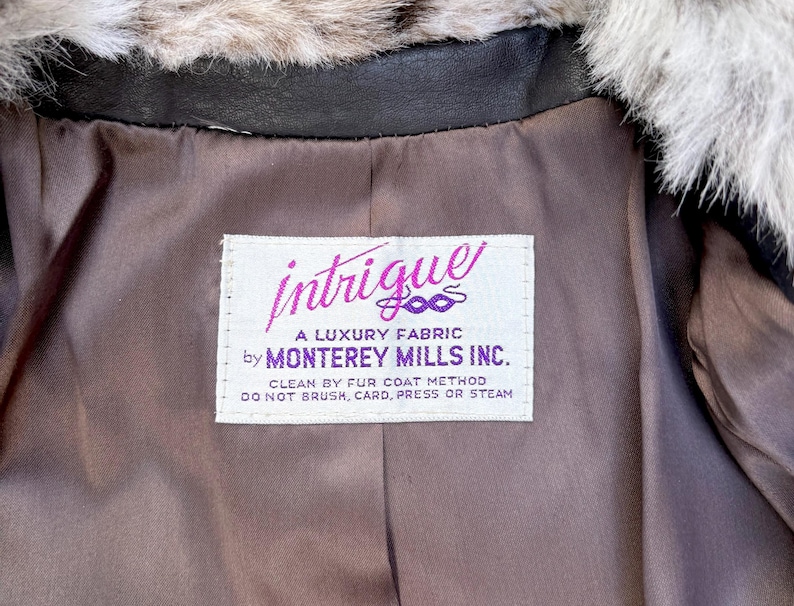 May include: A brown jacket interior with a white label reading intrigue A LUXURY FABRIC by MONTEREY MILLS INC. CLEAN BY FUR COAT METHOD DO NOT BRUSH, CARD, PRESS OR STEAM. The jacket features a dark brown lining and a fluffy grey and white faux fur collar.