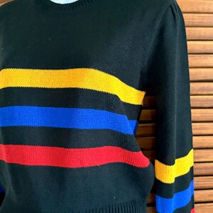 Pu&ograve; includere: Maglione nero con strisce orizzontali gialle, blu e rosse. Il maglione ha un girocollo e maniche lunghe. Le strisce sono uniformemente distanziate sul petto e sulle maniche.