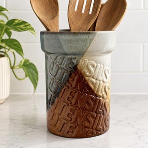 May include: A cylindrical ceramic utensil holder with a multi-toned glaze in shades of blue, white, and brown. The exterior features the word UTENSILS embossed repeatedly in a diagonal pattern. Three wooden kitchen utensils are placed inside.