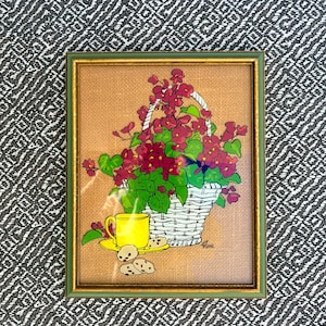 May include: Framed artwork featuring a still life scene. A white wicker basket overflows with red flowers and green leaves. A yellow teacup and saucer sit beside a plate of cookies. The frame is green with a gold inner border.