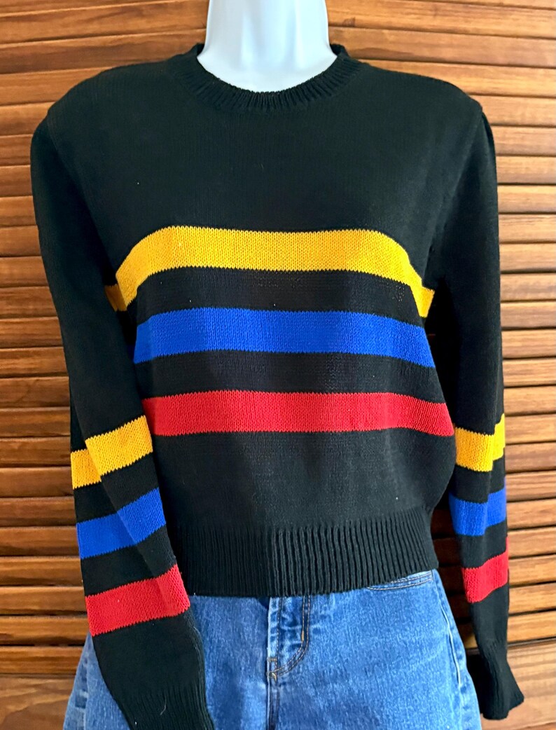 Pu&ograve; includere: Maglione nero con strisce orizzontali gialle, blu e rosse. Il maglione ha un girocollo e polsini e orlo a coste. Anche le maniche presentano il motivo a righe. Il maglione &egrave; esposto su un manichino.