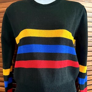 Pu&ograve; includere: Maglione nero con strisce orizzontali gialle, blu e rosse. Il maglione ha un girocollo e polsini e orlo a coste. Anche le maniche presentano delle strisce. Il maglione &egrave; esposto su un manichino.
