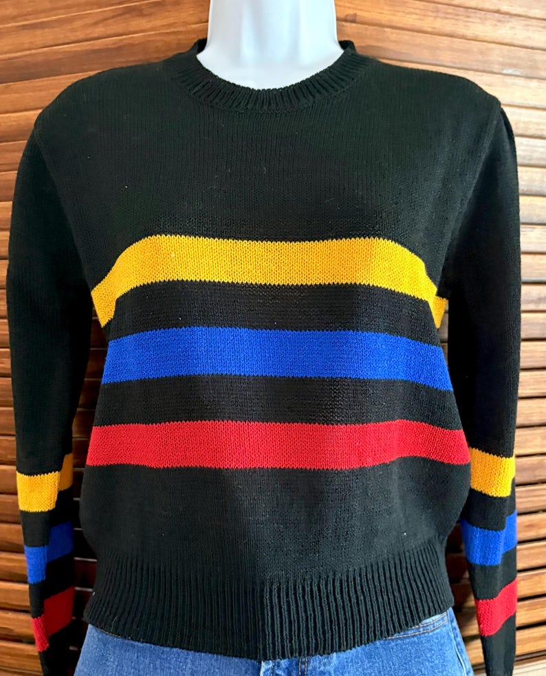 Pu&ograve; includere: Maglione nero con strisce orizzontali gialle, blu e rosse. Il maglione ha un girocollo e polsini e orlo a coste. Anche le maniche presentano delle strisce. Il maglione &egrave; esposto su un manichino.