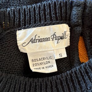 Pu&ograve; includere: Primo piano di un maglione nero a maglia con strisce blu e arancioni. L'etichetta recita "Adrianna Papell" e indica che il materiale &egrave; 80% acrilico e 20% nylon. Taglia S.