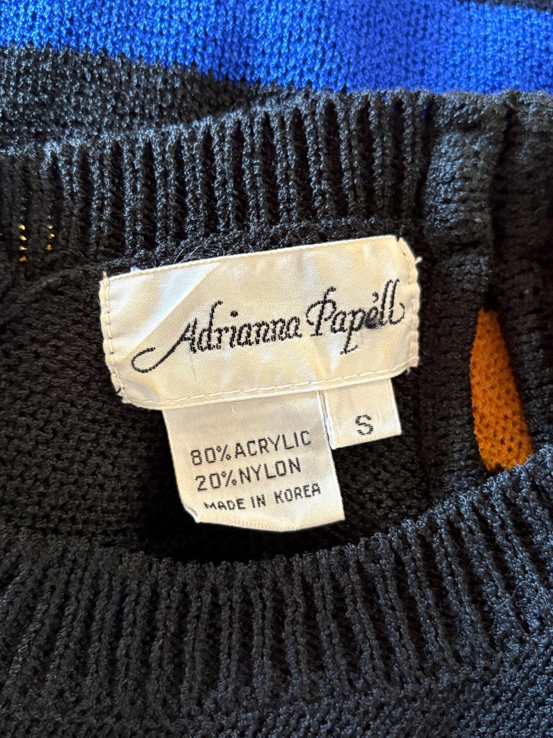 Pu&ograve; includere: Primo piano di un maglione nero a maglia con strisce blu e arancioni. L'etichetta recita "Adrianna Papell" e indica che il materiale &egrave; 80% acrilico e 20% nylon. Taglia S.