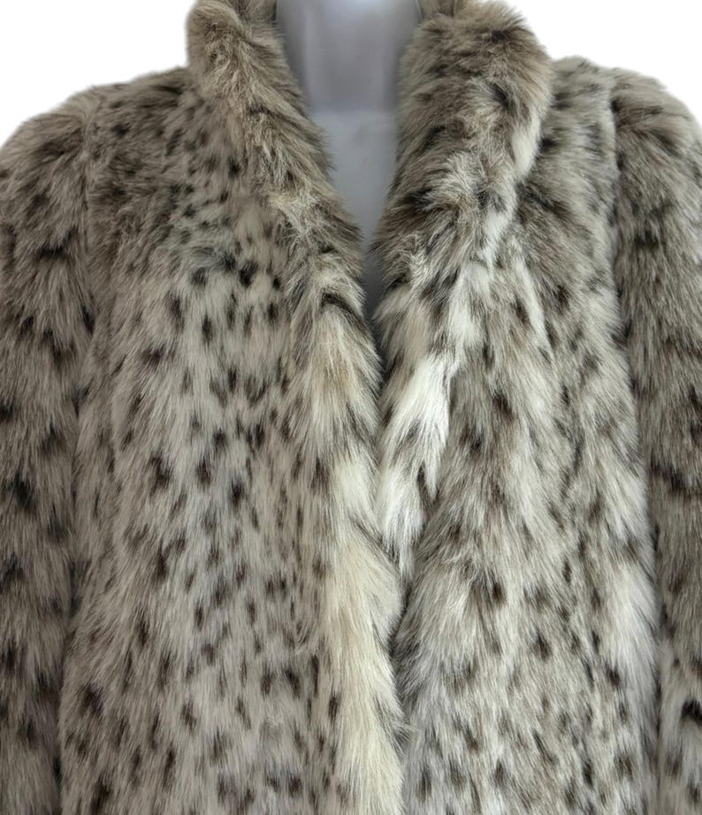 May include: A long-sleeved coat made of faux fur with a thick, plush collar. The garment features a light cream and beige base color with a dense pattern of dark brown and black spots distributed across the entire surface of the textured material.