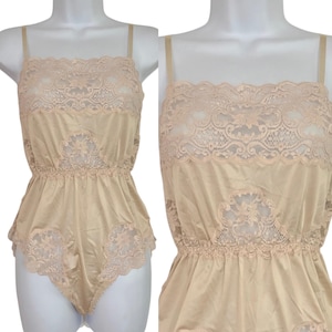 May include: A beige bodysuit featuring a square neckline with intricate lace detailing across the chest and waist. The garment has thin shoulder straps and a smooth, satin-like fabric finish, with additional lace trim along the leg openings.