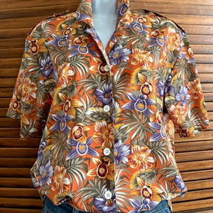 May include: Orange button-down shirt with a vibrant floral pattern featuring purple, yellow, and orange flowers, and green leaves. The short-sleeved shirt has a collared neckline and white buttons. The shirt is displayed on a mannequin.
