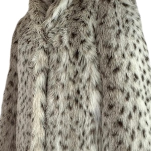 May include: A long-sleeved coat featuring a dense, textured faux fur exterior with a cream-colored base and a repeating pattern of dark brown and black spots throughout the garment. The coat has a high collar and a straight, structured silhouette.