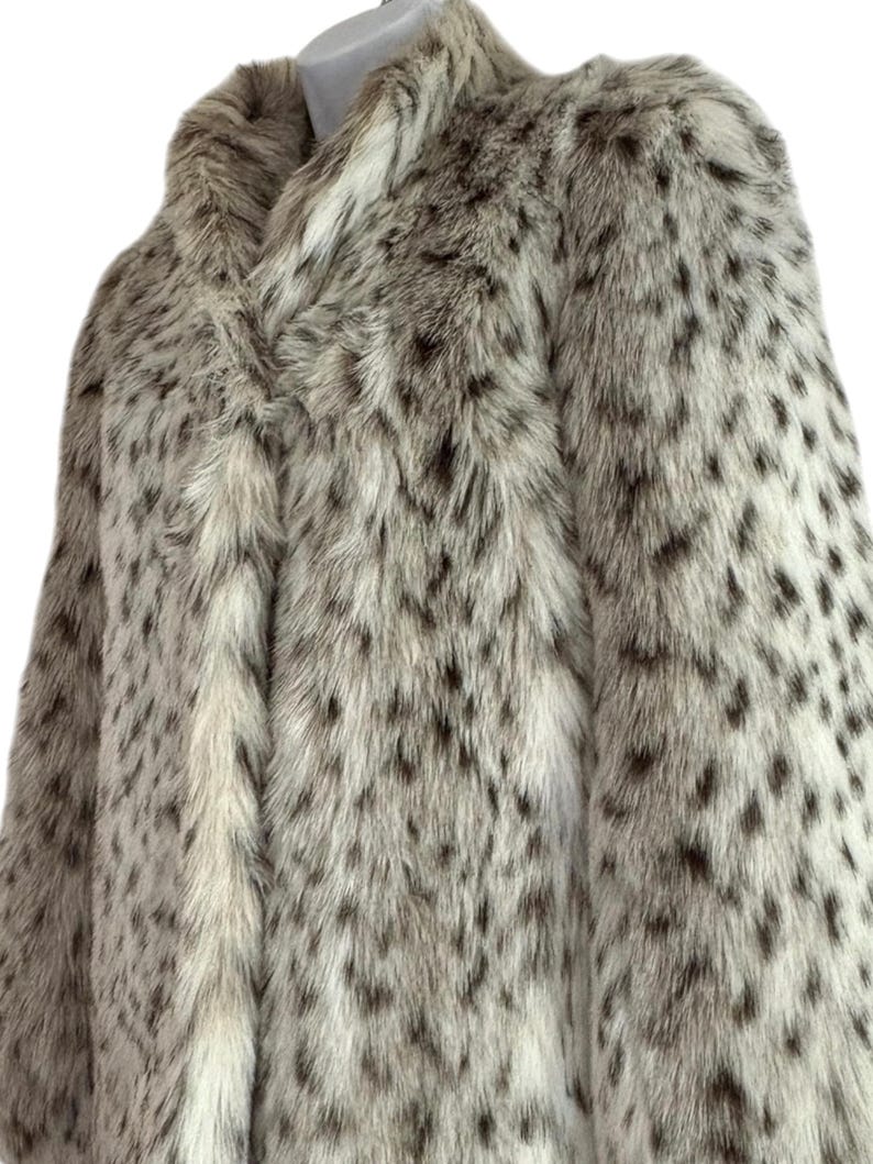 May include: A long-sleeved coat featuring a dense, textured faux fur exterior with a cream-colored base and a repeating pattern of dark brown and black spots throughout the garment. The coat has a high collar and a straight, structured silhouette.
