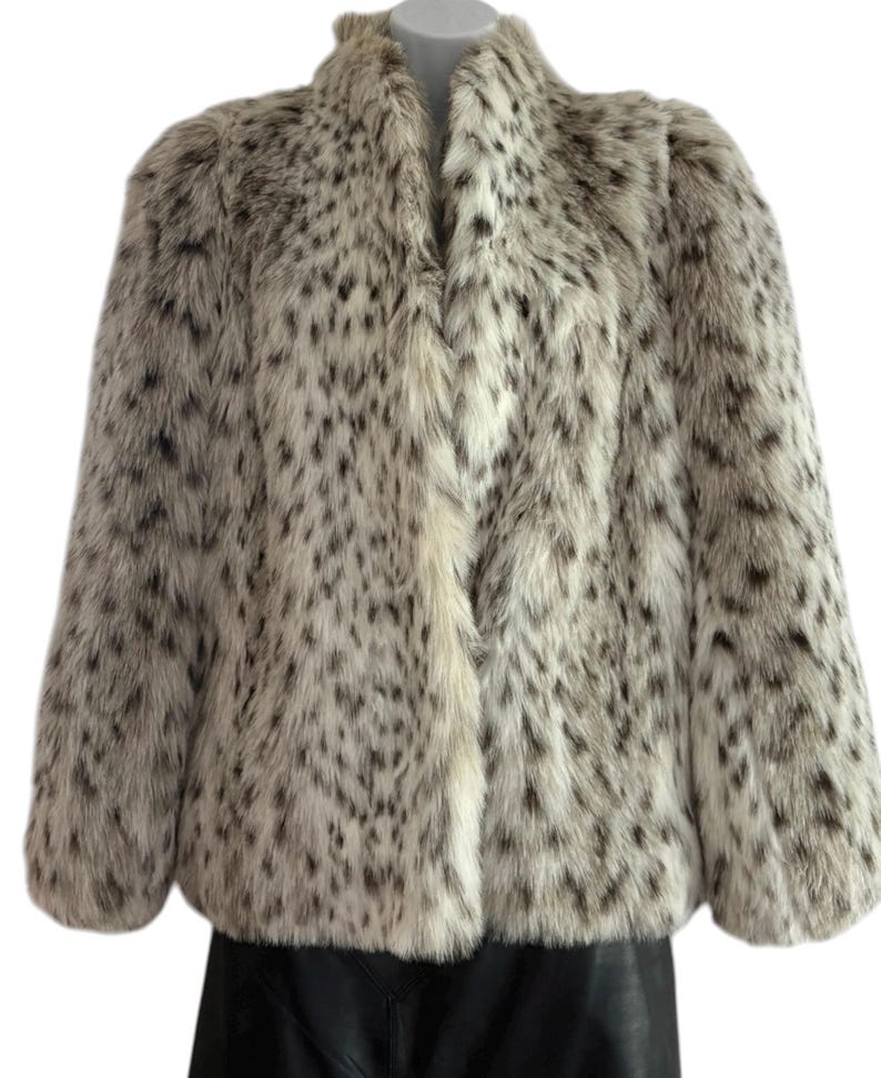 May include: A long-sleeved faux fur jacket featuring a cream-colored base with a dense pattern of dark brown and black spots. The jacket has a high collar, a straight hemline, and a front opening that extends from the neck to the bottom edge.