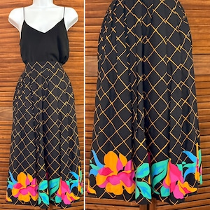 May include: Black dress with a V-neck top and a pleated skirt. The skirt features a gold diamond grid pattern and a vibrant floral border with pink, orange, and blue flowers. The dress is displayed on a mannequin.